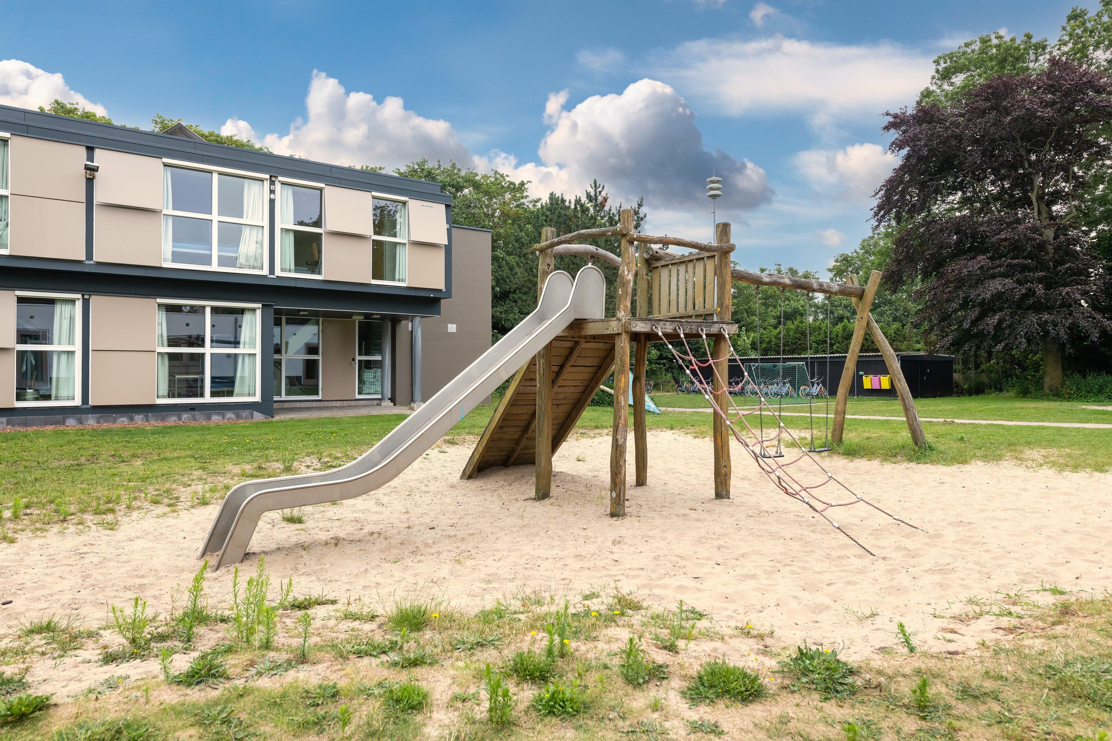 Ein Spielplatz mit Rutsche und Klettergerüsten, vor einer Gebäudeecke mit großen Fenstern und blauem Himmel.