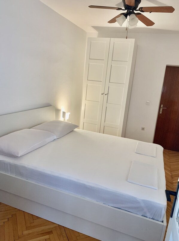 A neatly made bed with white linens, a ceiling fan, a wardrobe, and a wooden floor.