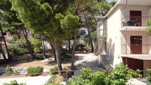 Um edifício residencial com escadaria que leva a uma varanda, rodeado por árvores e um muro de pedra.
