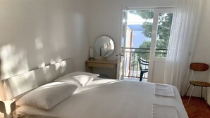 Cama bem arrumada com almofadas e mesa de cabeceira com abajur e espelho. Há uma cadeira e uma varanda com vista para árvores e um corpo de água.