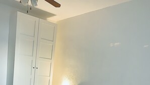 Quarto com cama, mesa de cabeceira, guarda-roupa e ventoinha no teto.