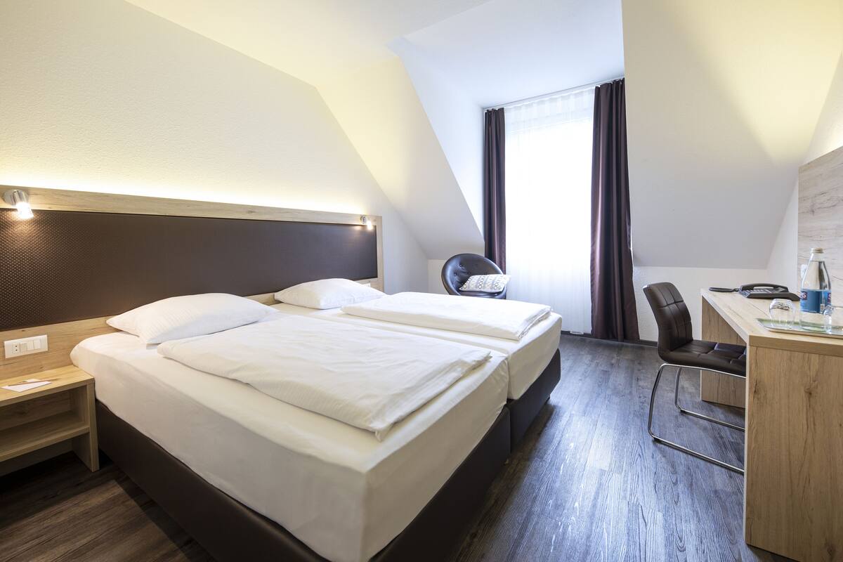 Habitación de hotel con dos camas, un escritorio, una silla y un ventanal con cortinas.