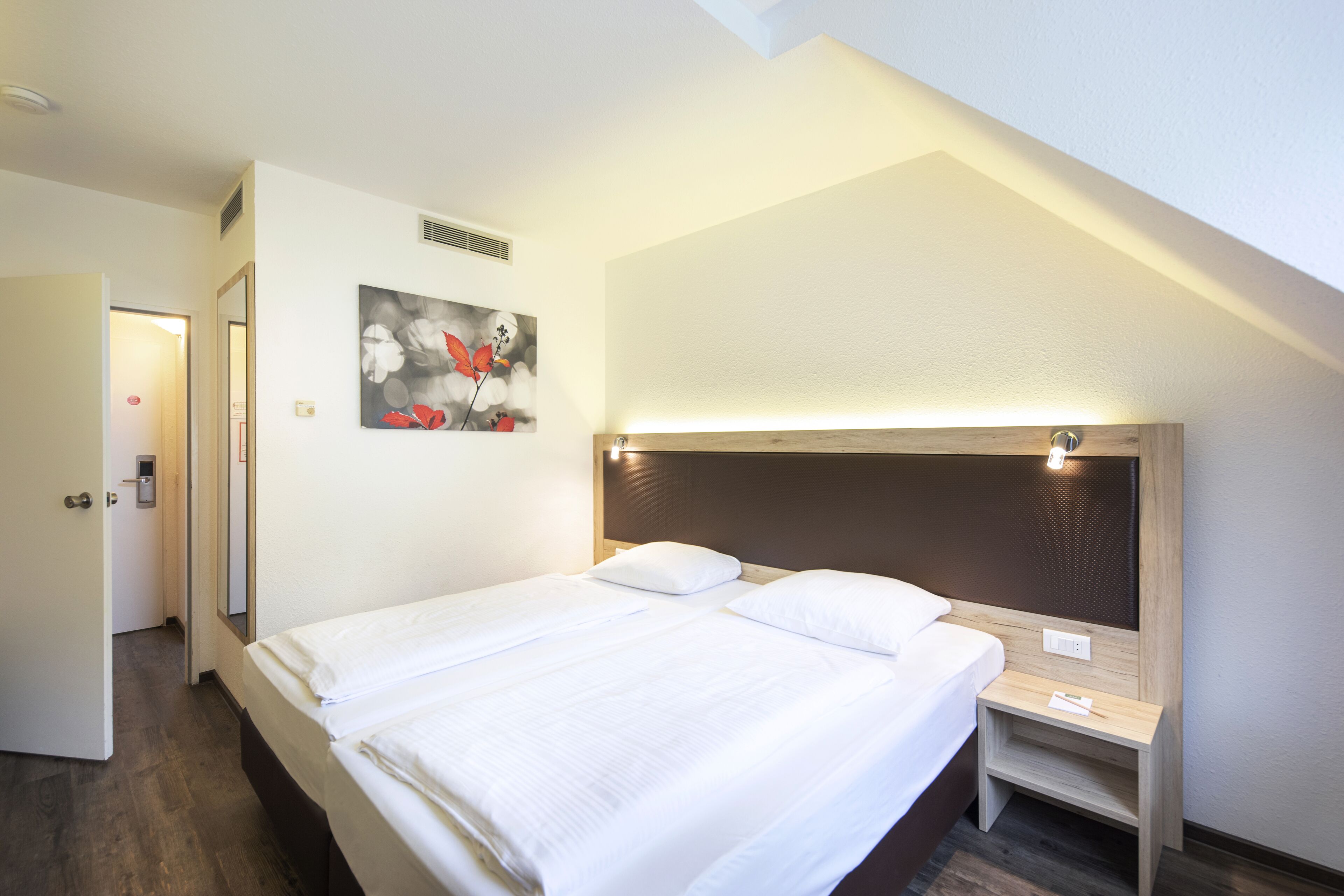 A hotel room with a neatly made bed, a bedside table, a nightstand, a wall-mounted artwork, and a door leading to another room.