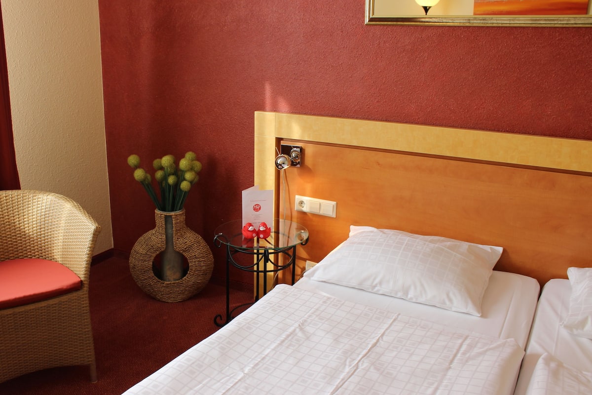 Una habitación de hotel con una cama, un sillón de mimbre con un cojín rojo, una mesita con un florero de flores verdes y un interruptor de luz en la pared.