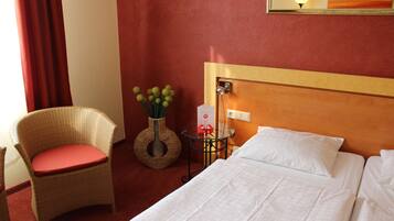 Een hotelkamer met een bed, een rieten stoel met een rood kussen, een kleine tafel met een vaas groene bloemen en een wandlampen schakelaar.