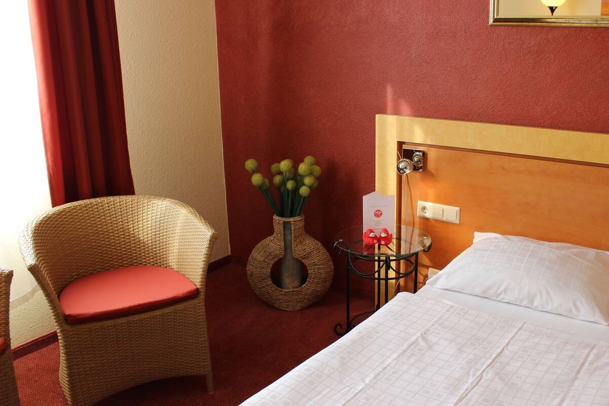 Una habitación de hotel con una cama, una silla de mimbre con un cojín rojo, un florero con flores verdes y una mesita con una lámpara.