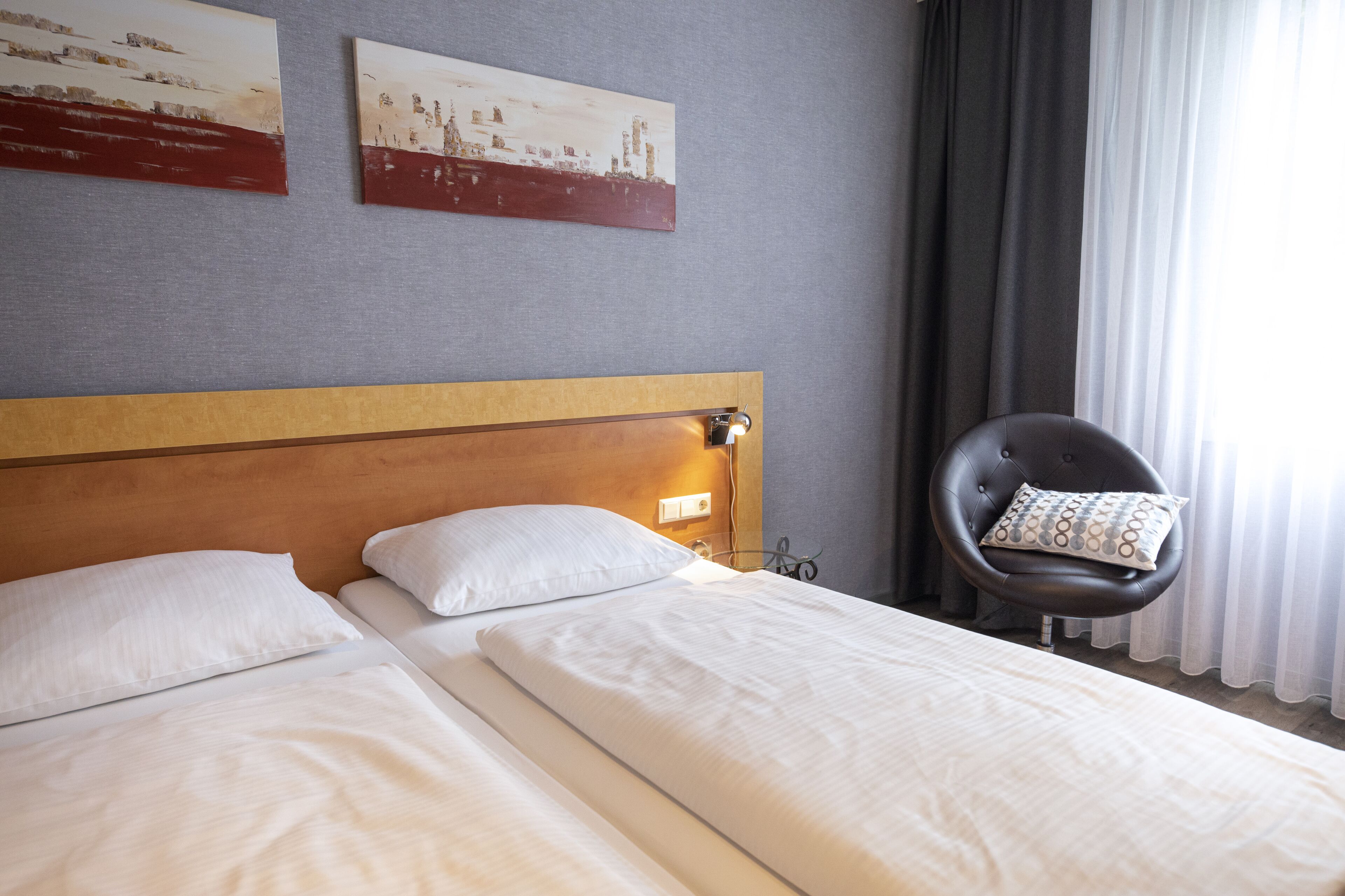 Una camera d'albergo con due letti singoli, una testiera in legno, una sedia nera con cuscino decorato e due quadri appesi al muro.