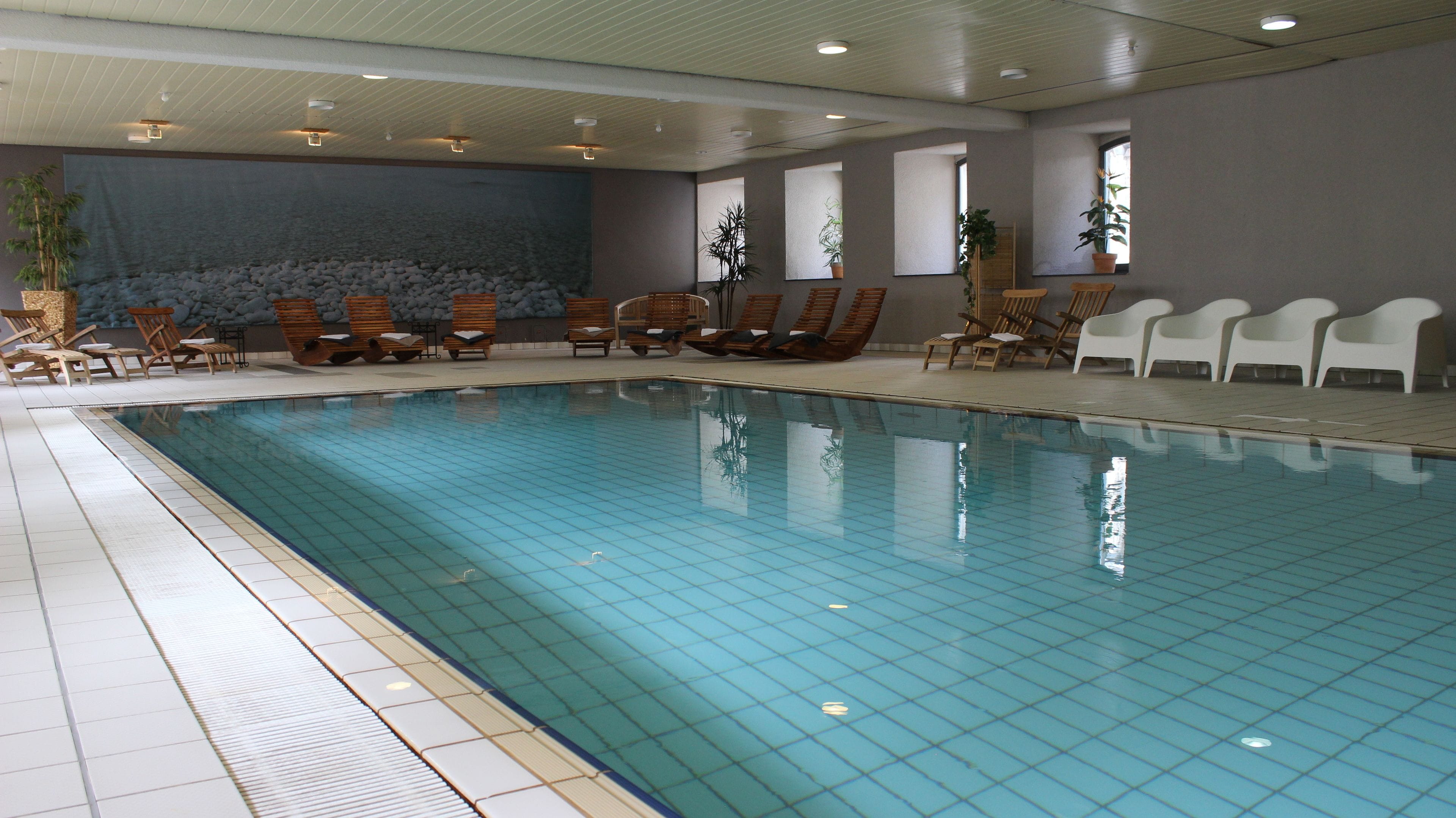 A swimming pool with tiled flooring, wooden lounge chairs, and potted plants.