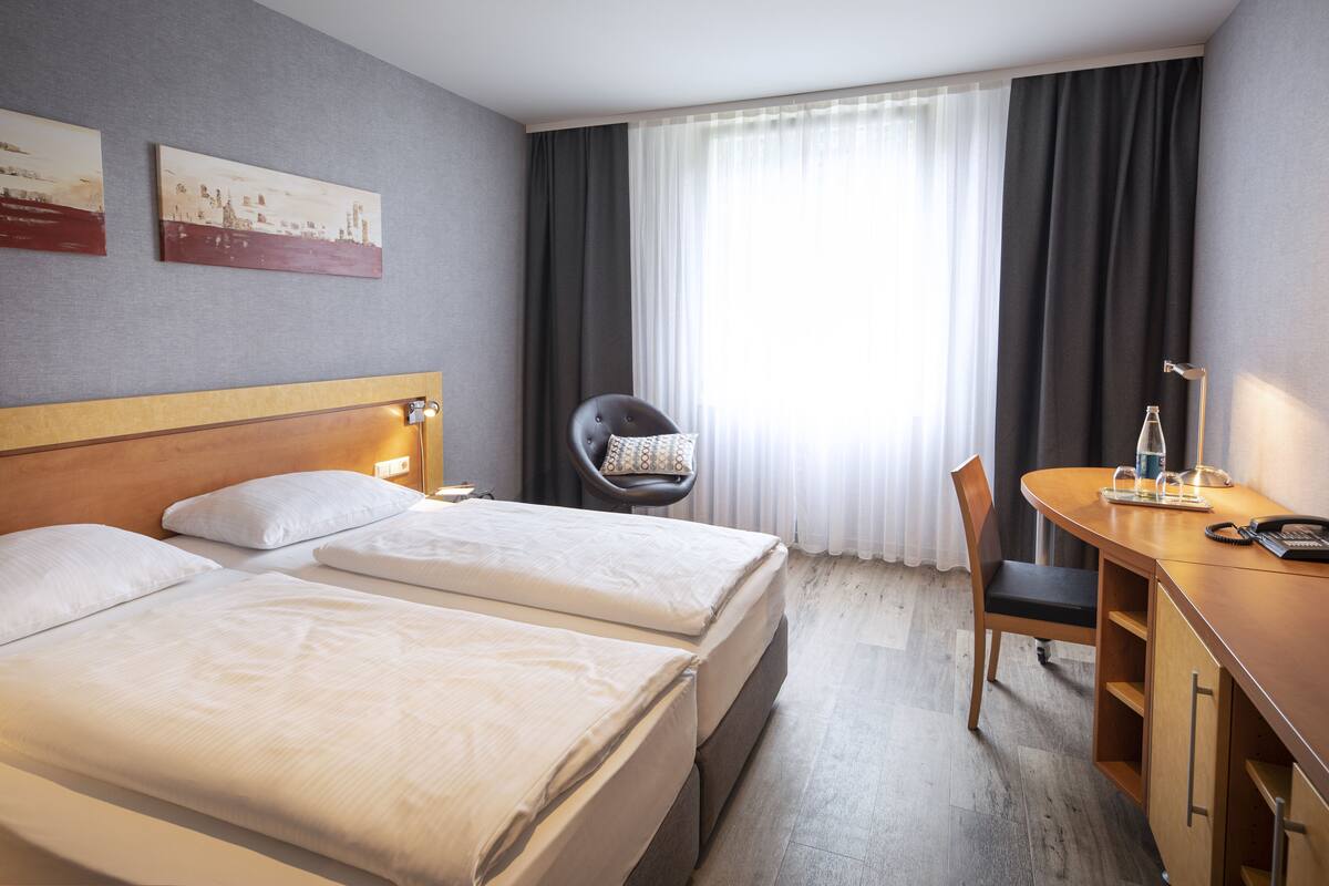 Una habitación de hotel con dos camas, un escritorio, una silla y un ventanal con cortinas.
