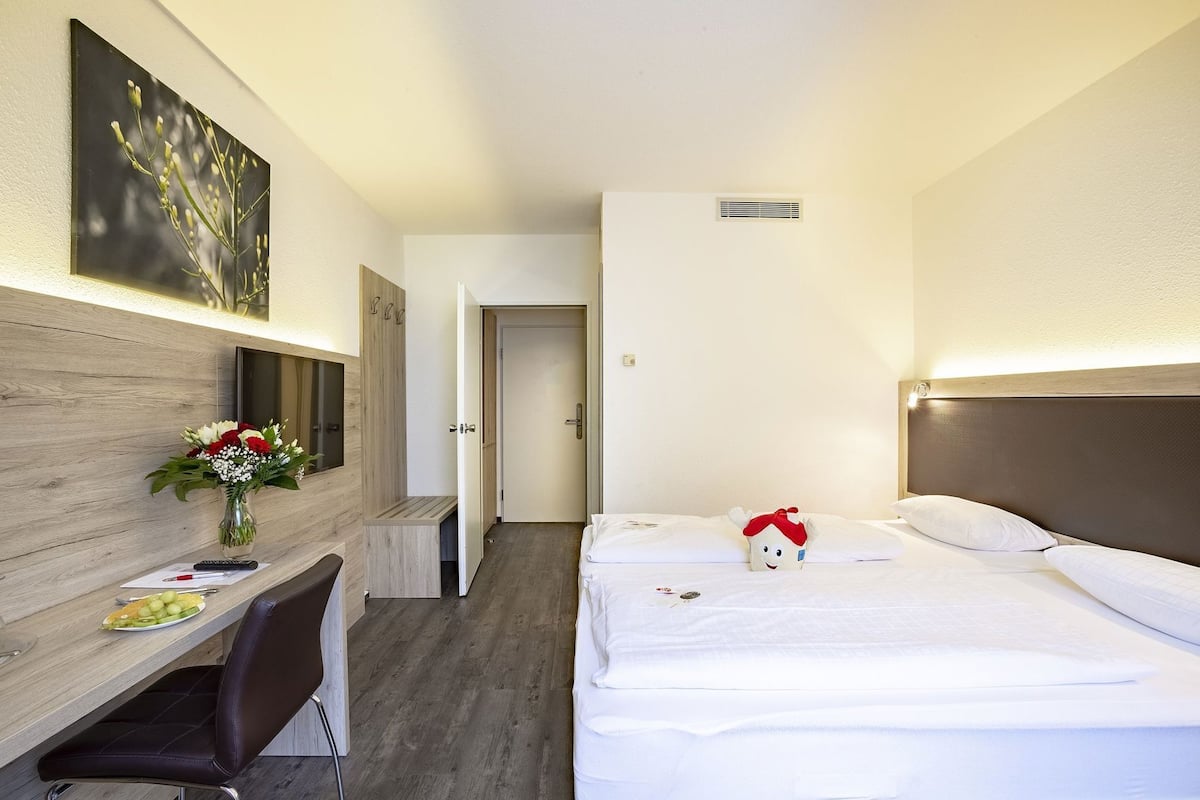 Habitación de hotel con cama, escritorio y silla, un jarrón de flores y un cuadro en la pared.