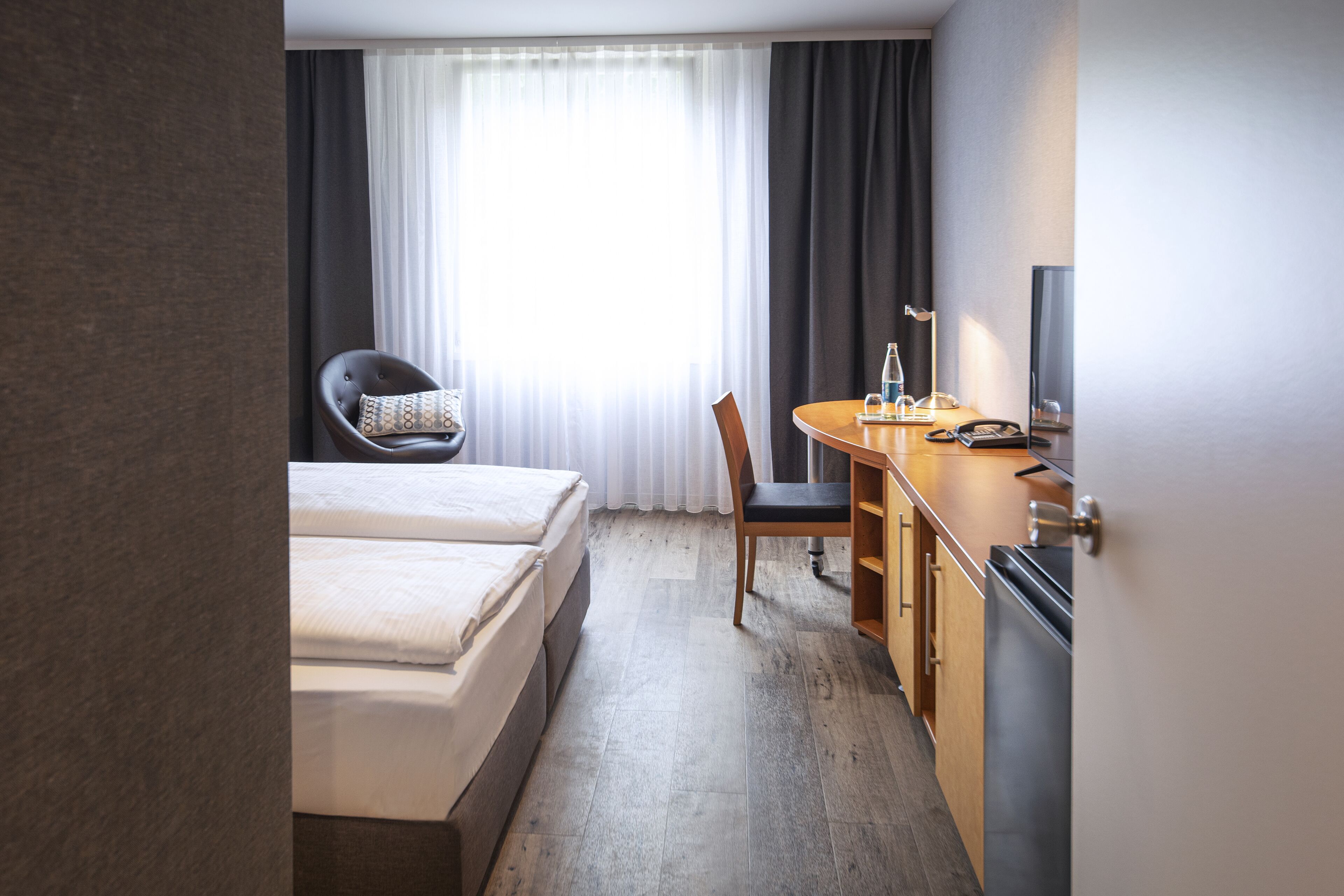 A hotel room with a bed, a desk with a chair, a television, and a window with curtains.
