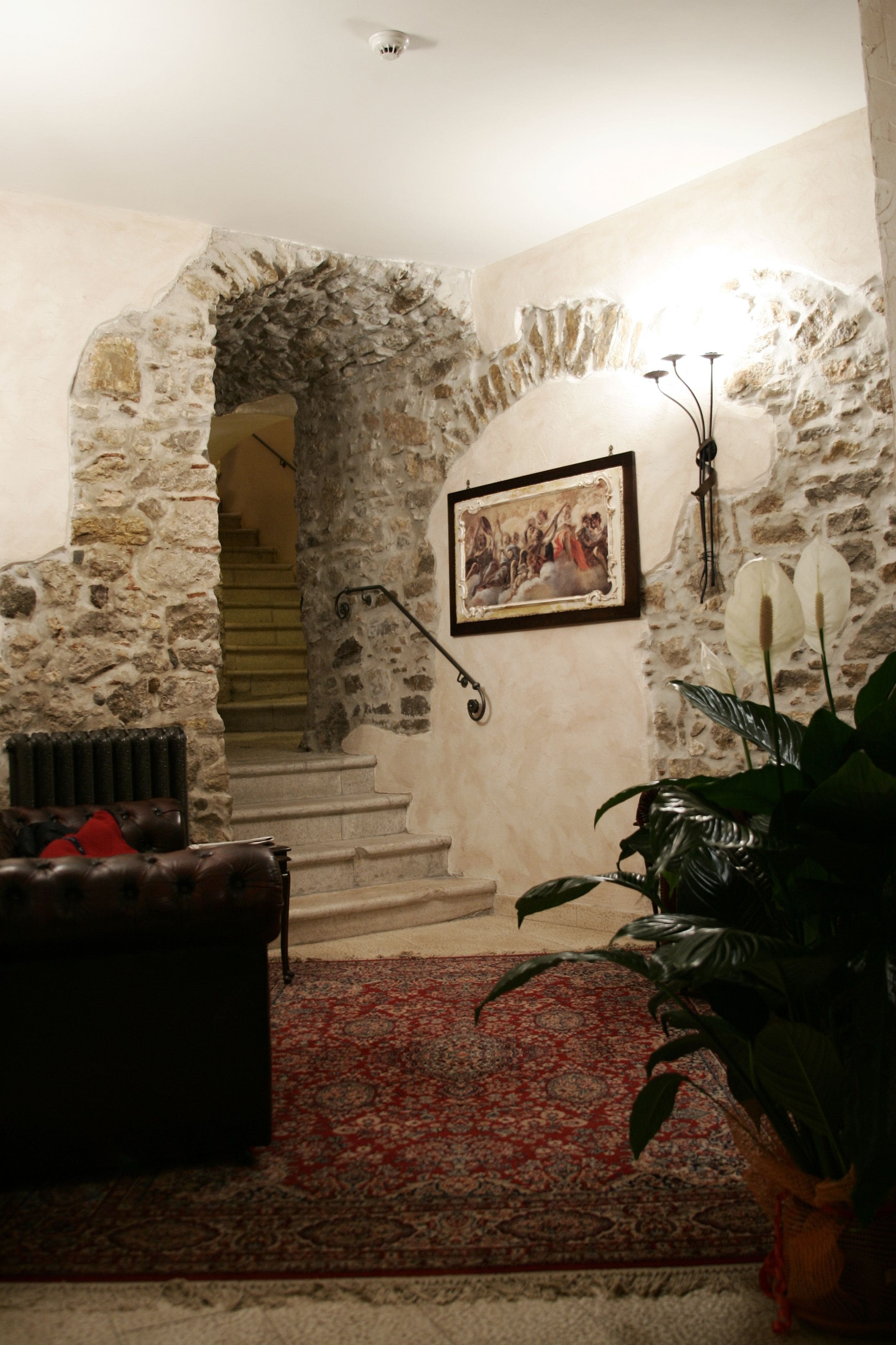 A stone-walled room with a staircase, a framed picture, a radiator, a leather sofa, and a potted plant.