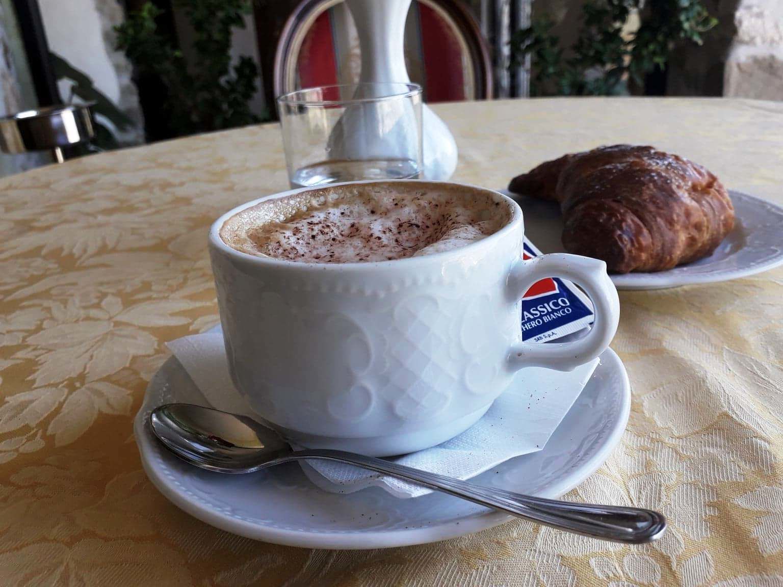 A cup of coffee with a spoon on a saucer, a croissant, and a glass of water on a table with a patterned tablecloth.