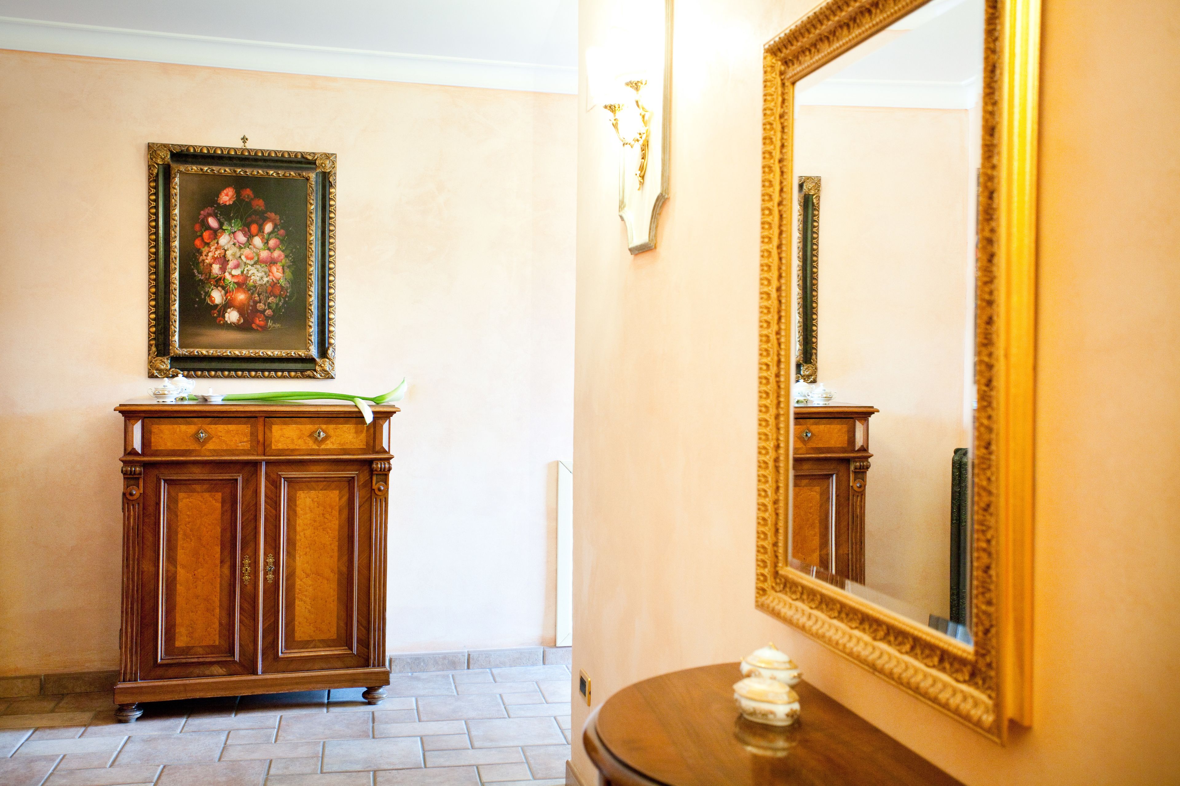 A hallway with a wooden cabinet, a framed floral painting, and a large mirror on the wall.