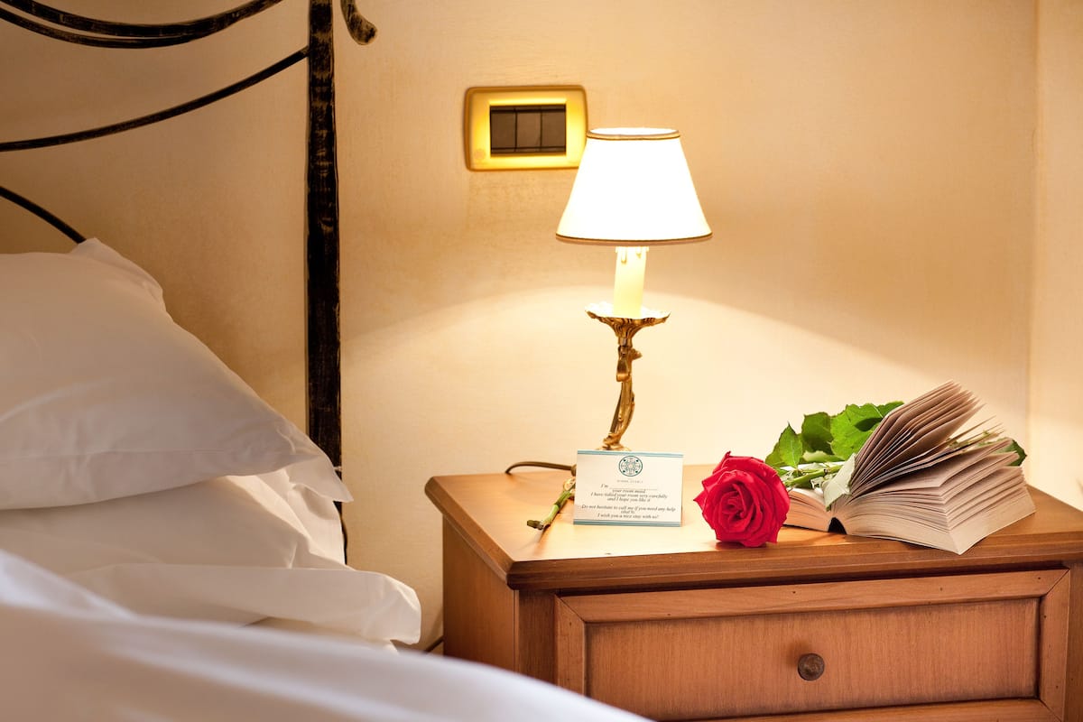 A neatly made bed with white linens, a bedside lamp with a wooden base, a red rose, and an open book on a wooden nightstand.