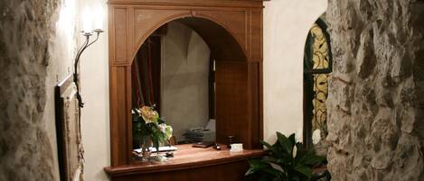 Interior com arco de pedra que leva a um quarto com uma escrivaninha de madeira, um espelho, um vaso de planta e um tapete estampado.