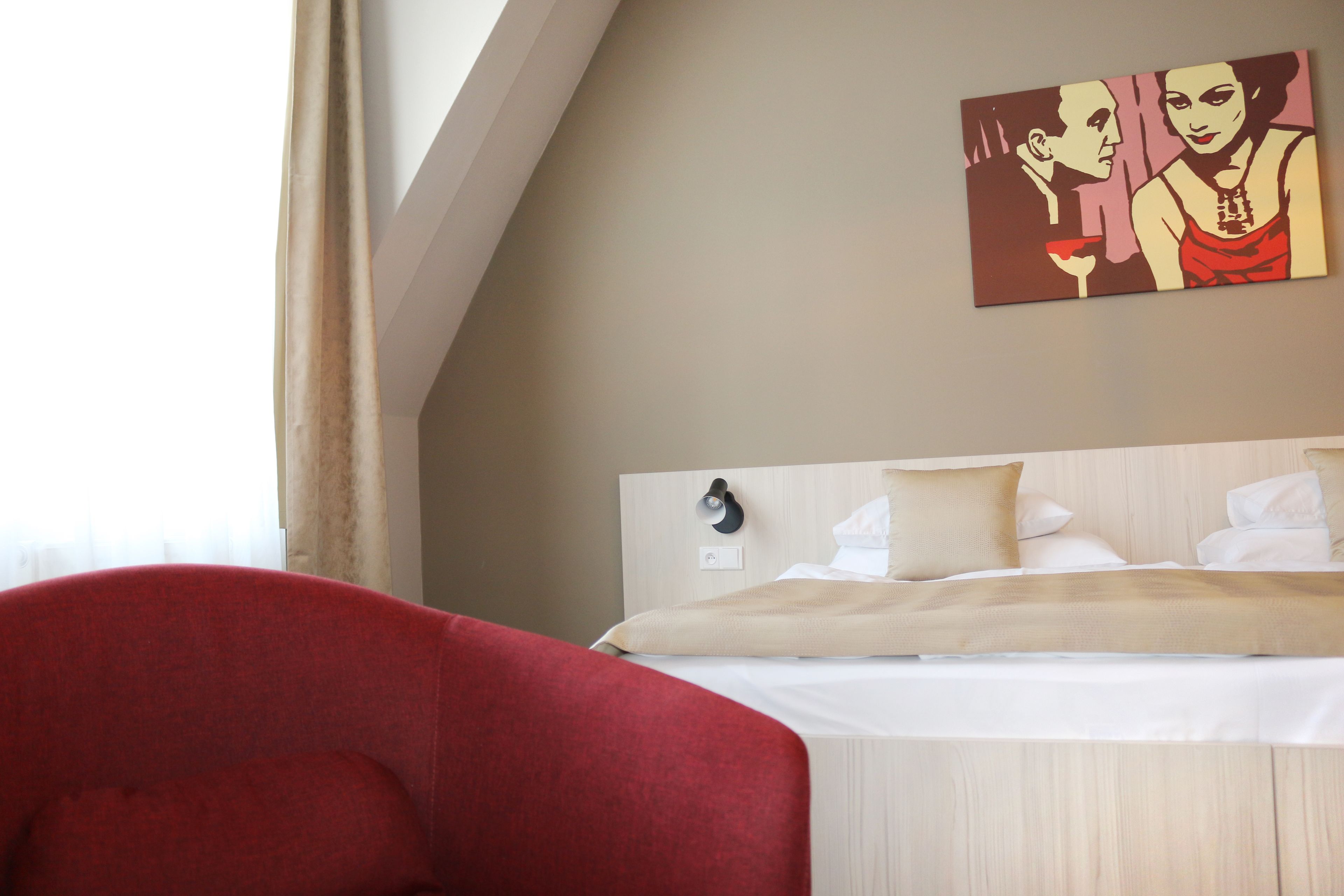 Une chambre d’hôtel avec un lit, un fauteuil rouge, un tableau au mur et une fenêtre avec des rideaux.