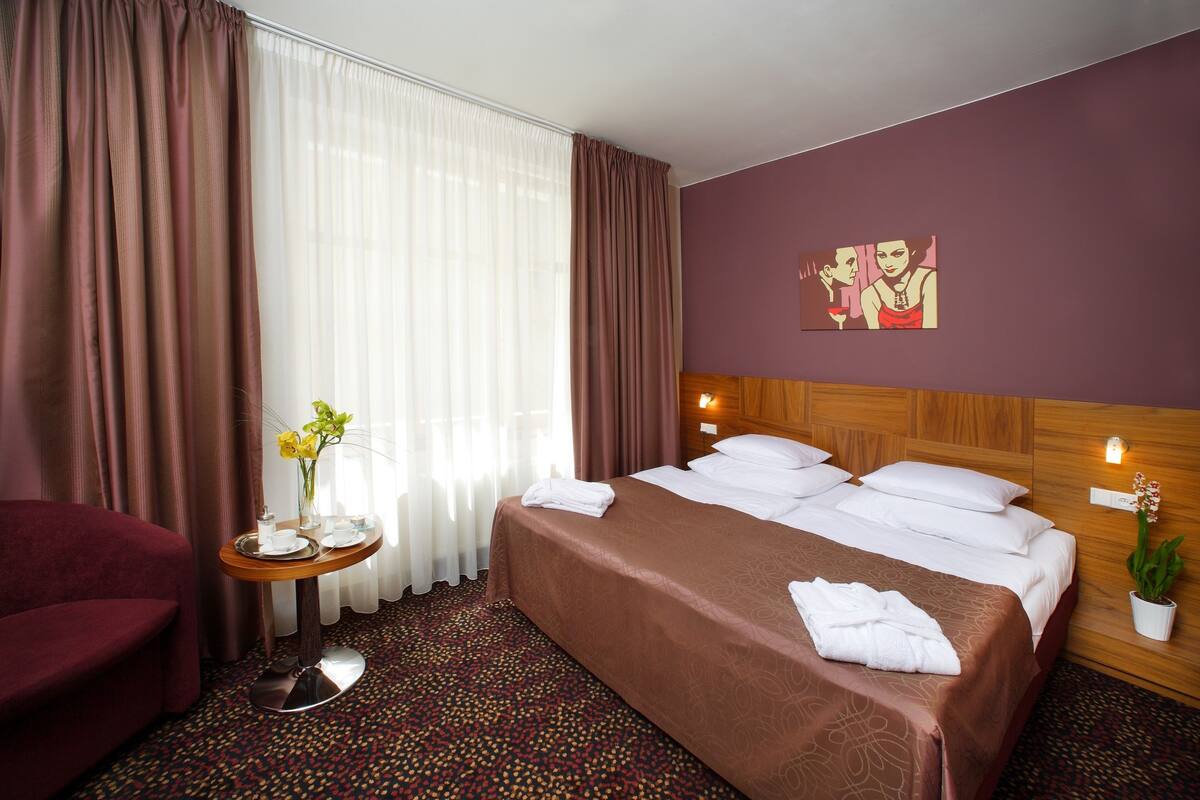 A hotel room with a large bed, a wooden headboard, a small table with a vase of flowers, and a red armchair.