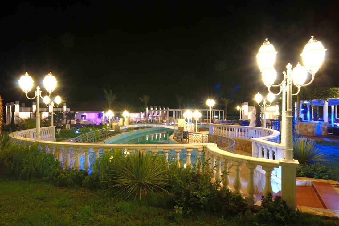 Vue nocturne d'un espace piscine agrémenté de lampadaires décoratifs et d'une fontaine, le tout entouré d'une balustrade blanche.