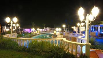 A nighttime view of a pool area with ornate lamp posts and a fountain, surrounded by a white balustrade.