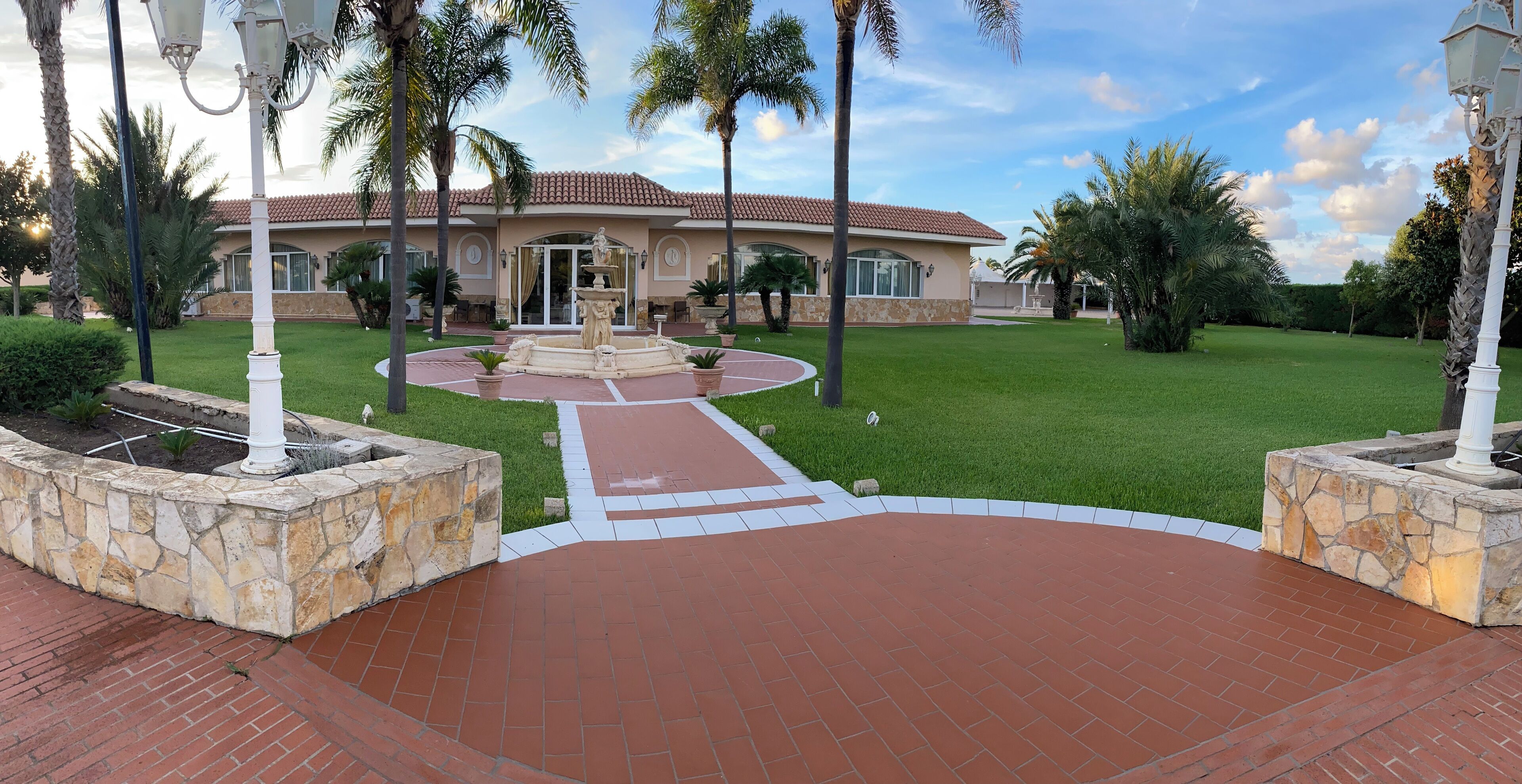 A well-maintained garden with a fountain, stone walls, and a pathway leading to a house with a tiled roof.
