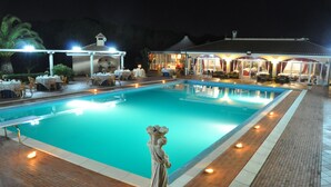 A swimming pool with a statue in the center, surrounded by tables and chairs under a covered area. - Agriturismo Al Parco (Lecce)