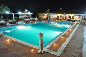 Una piscina con una estatua en el centro, rodeada de mesas y sillas bajo una zona cubierta.