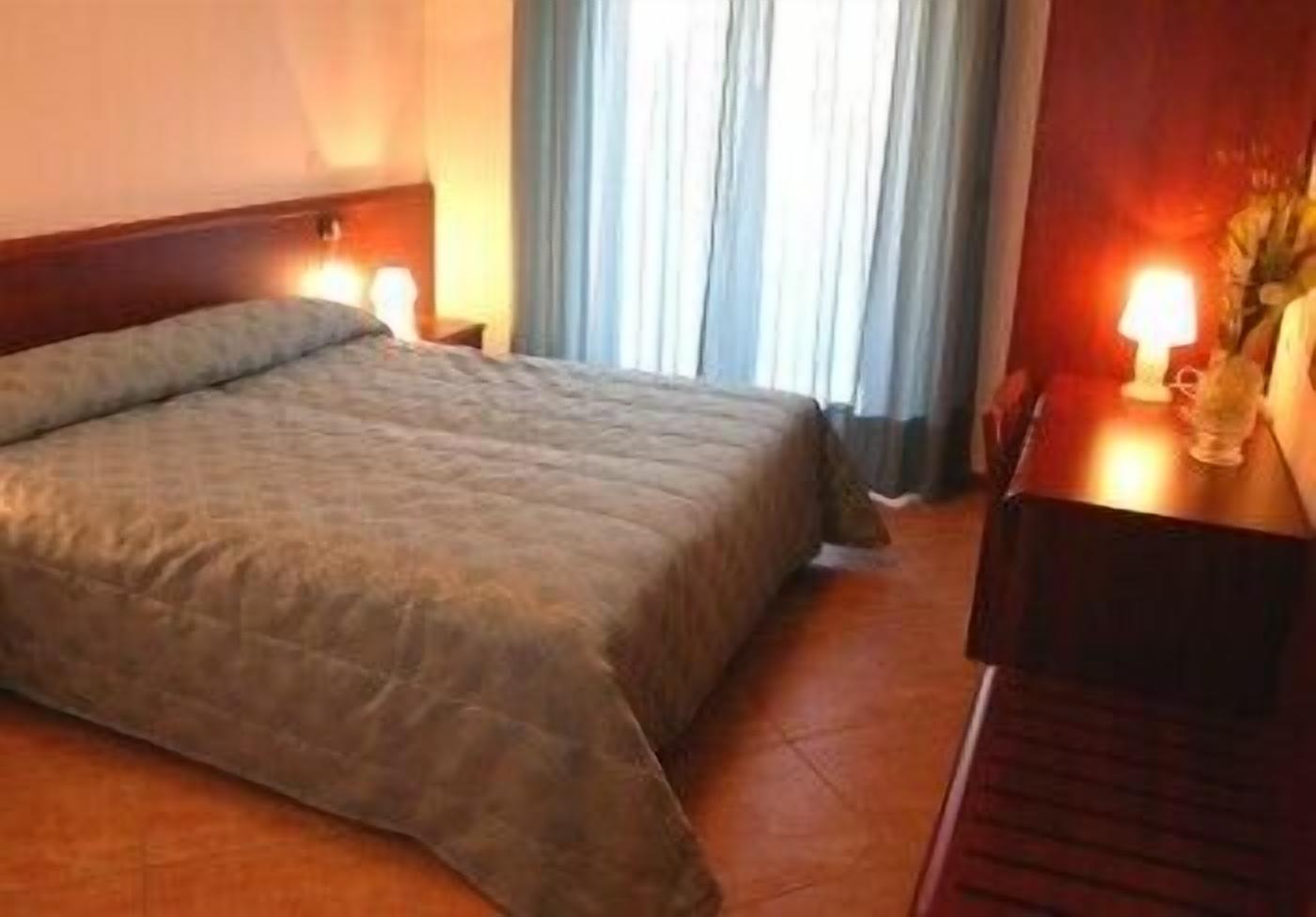 A hotel room with a large bed, a wooden headboard, a nightstand with a lamp, and a window with curtains.