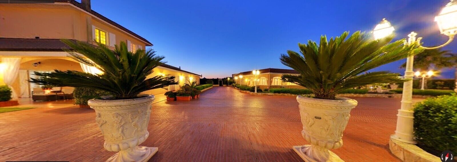 A hotel entrance with a paved pathway, potted plants, and illuminated street lamps.