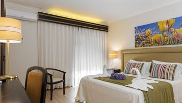 Quarto de hotel com uma cama, uma escrivaninha com abajur, uma cadeira e um quadro na parede.