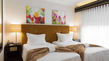 Habitación de hotel con dos camas, un cabecero de madera, dos mesitas de noche con lámparas y dos cuadros con motivos florales en la pared.