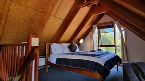 Une chambre mansardée chaleureuse avec un plafond en pente en bois, un lit recouvert d’une couvre-lit à rayures foncées et claires, une lampe de chevet et une fenêtre avec des rideaux.