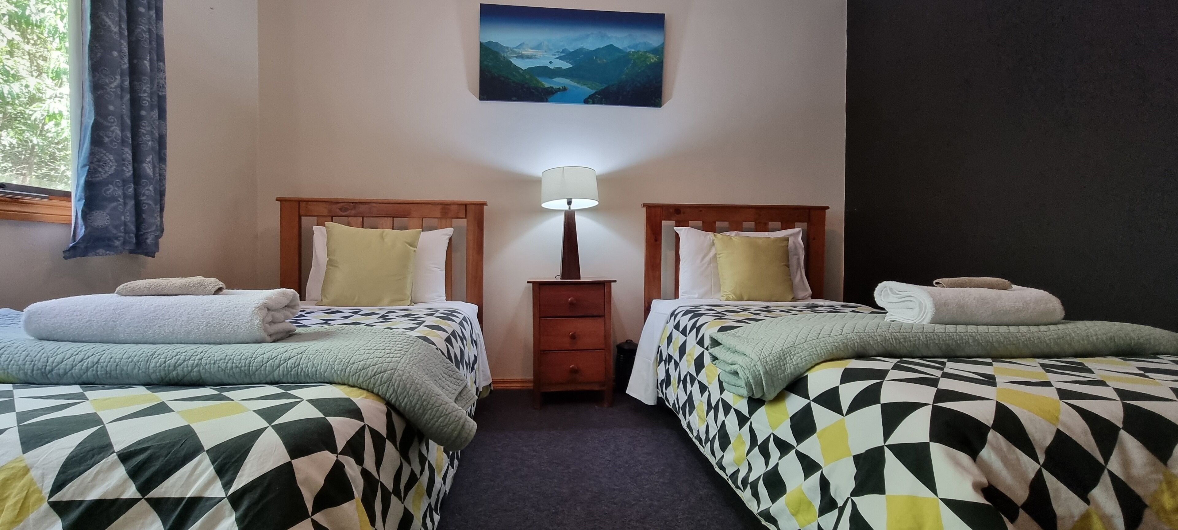 A room with two beds, each with a patterned bedspread and pillows, a nightstand with a lamp, and a painting on the wall.