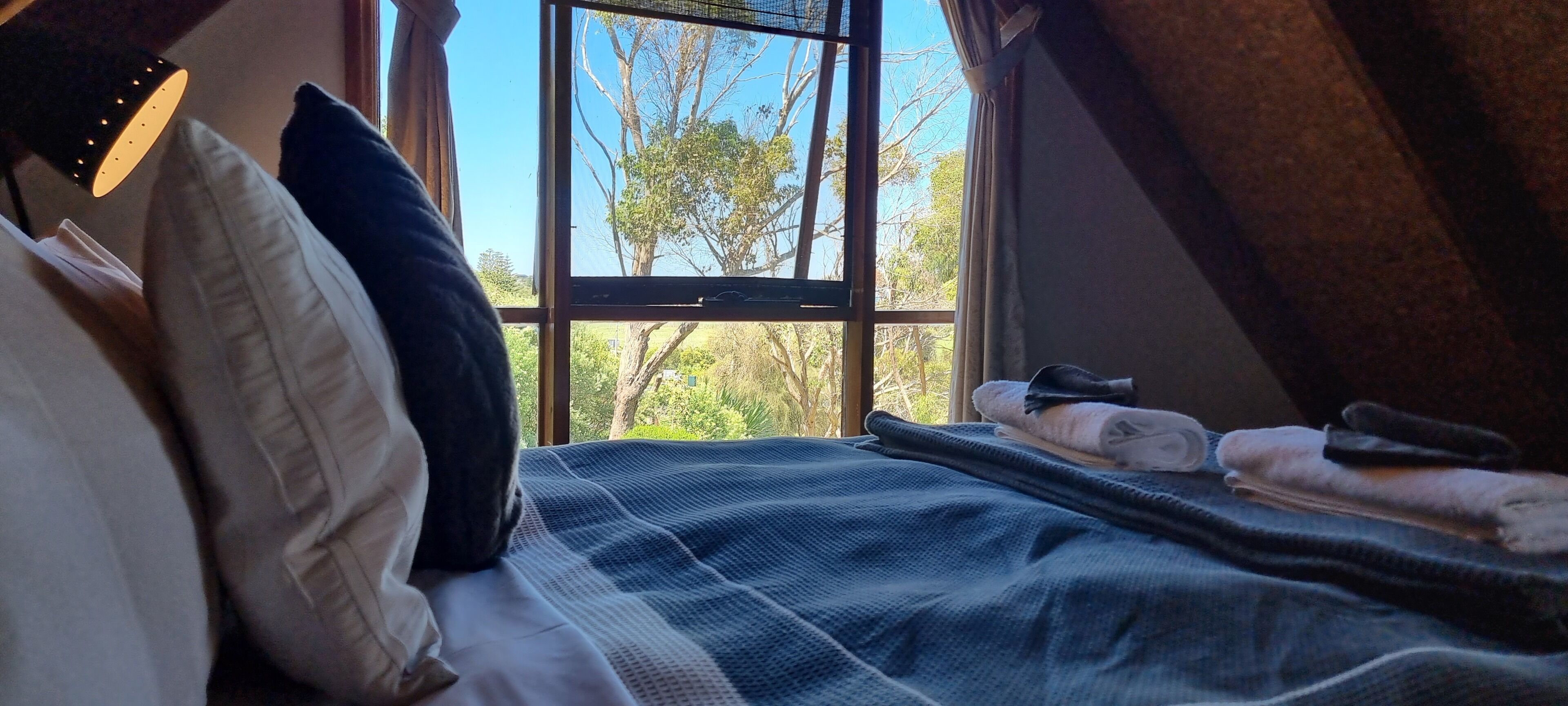 Une chambre avec une grande fenêtre, un lit recouvert d’une couverture à carreaux bleus et blancs, et une vue sur les arbres et la verdure à l’extérieur.