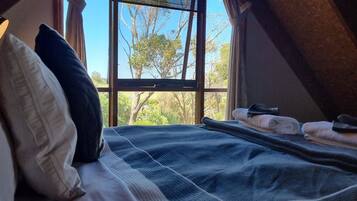 Une chambre avec une grande fenêtre, un lit recouvert d’une couverture à carreaux bleus et blancs, et une vue sur les arbres et la verdure à l’extérieur.