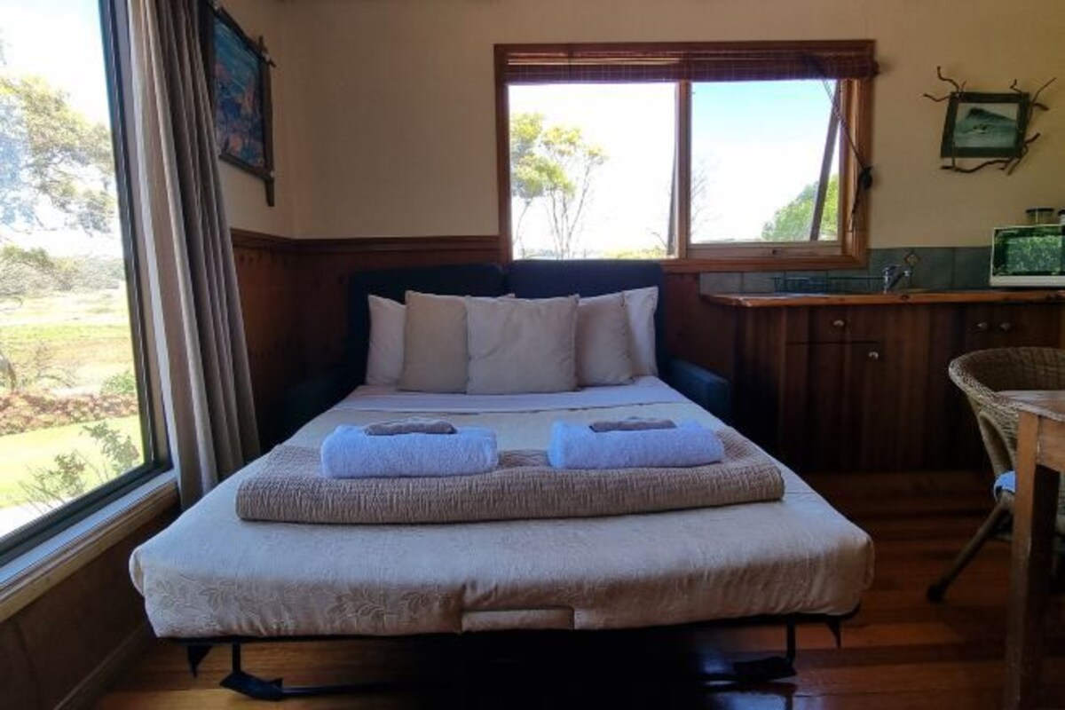 A single bed with a headboard, a window with blinds, a small kitchenette with a sink and a television, and a view of the outdoors.