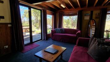 A cozy room with a wooden ceiling, a purple sofa, a wooden table with a laptop, and large windows offering a view of the outdoors.