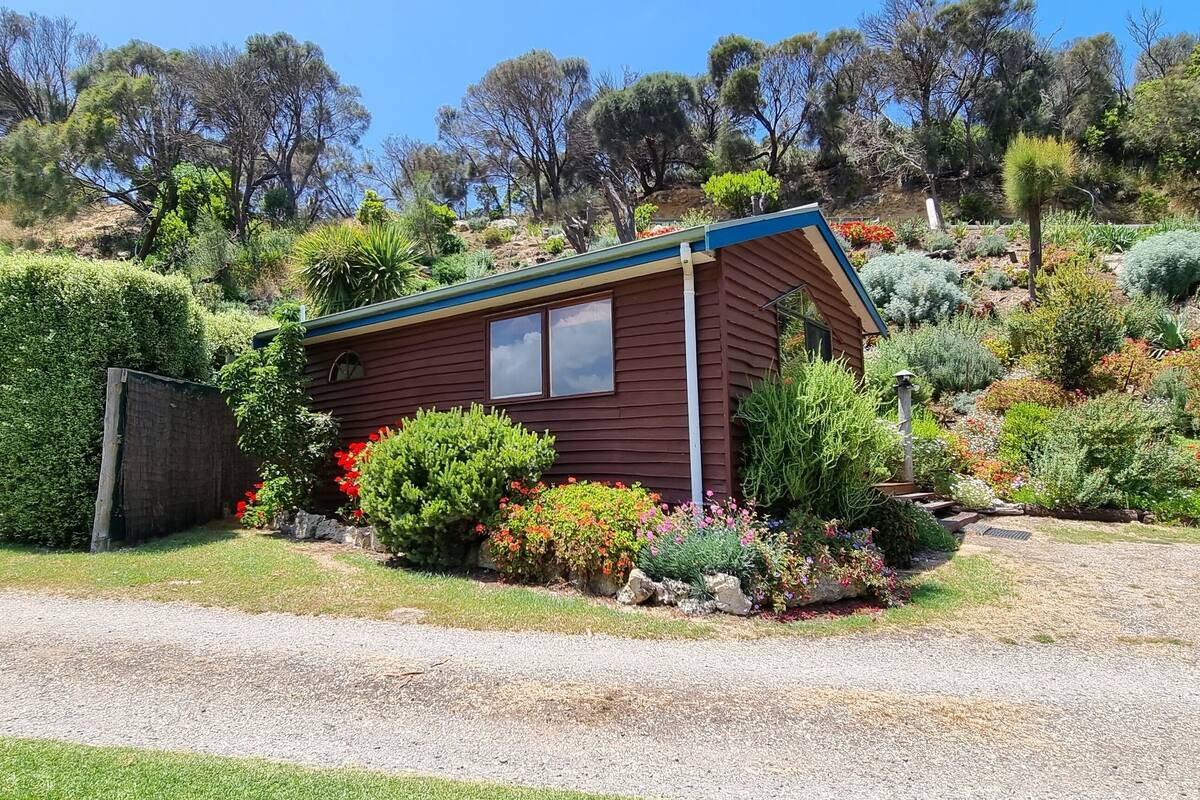 A small, single-story wooden house with a sloped roof, surrounded by a well-maintained garden with various plants and flowers.