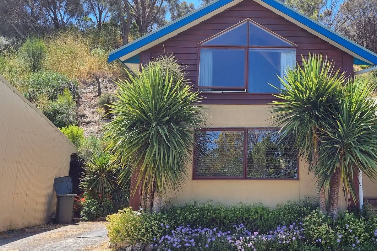A two-story house with a brown roof and blue trim, surrounded by lush greenery and flowering plants.