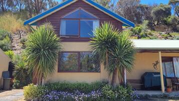Una casa de dos pisos con techo marrón y detalles azules, rodeada de exuberante follaje y plantas en flor.