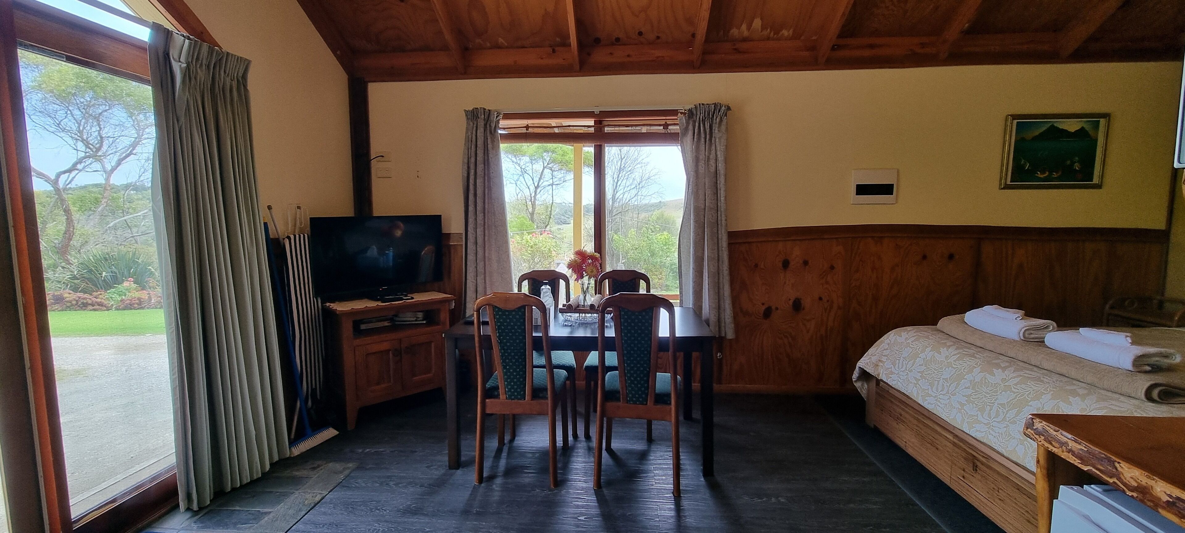 A room with a wooden ceiling, a TV on a wooden stand, a dining table with chairs, a bed with towels, and a window with curtains.