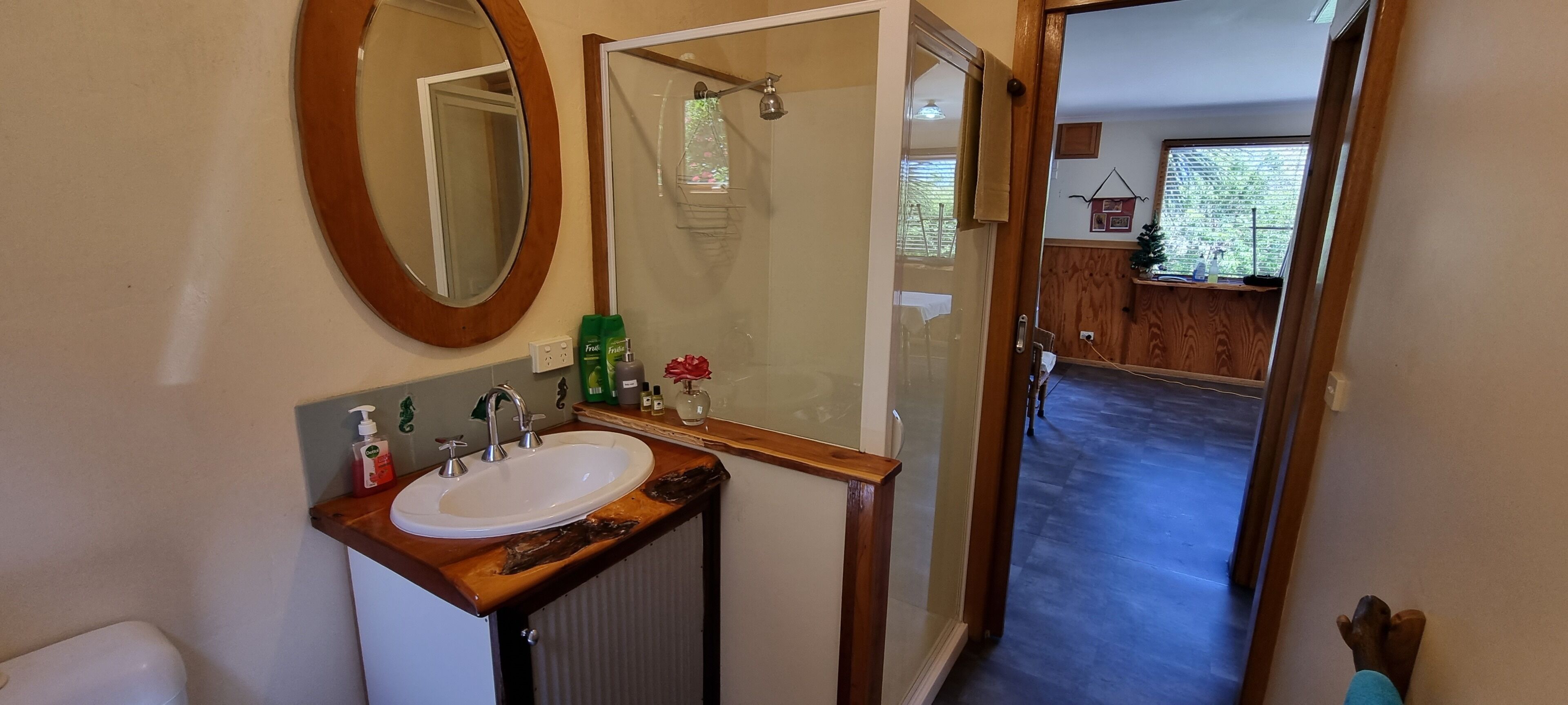 A bathroom with a wooden vanity, a white sink, and two mirrors. There are toiletries on the vanity and a shower visible through a glass door.