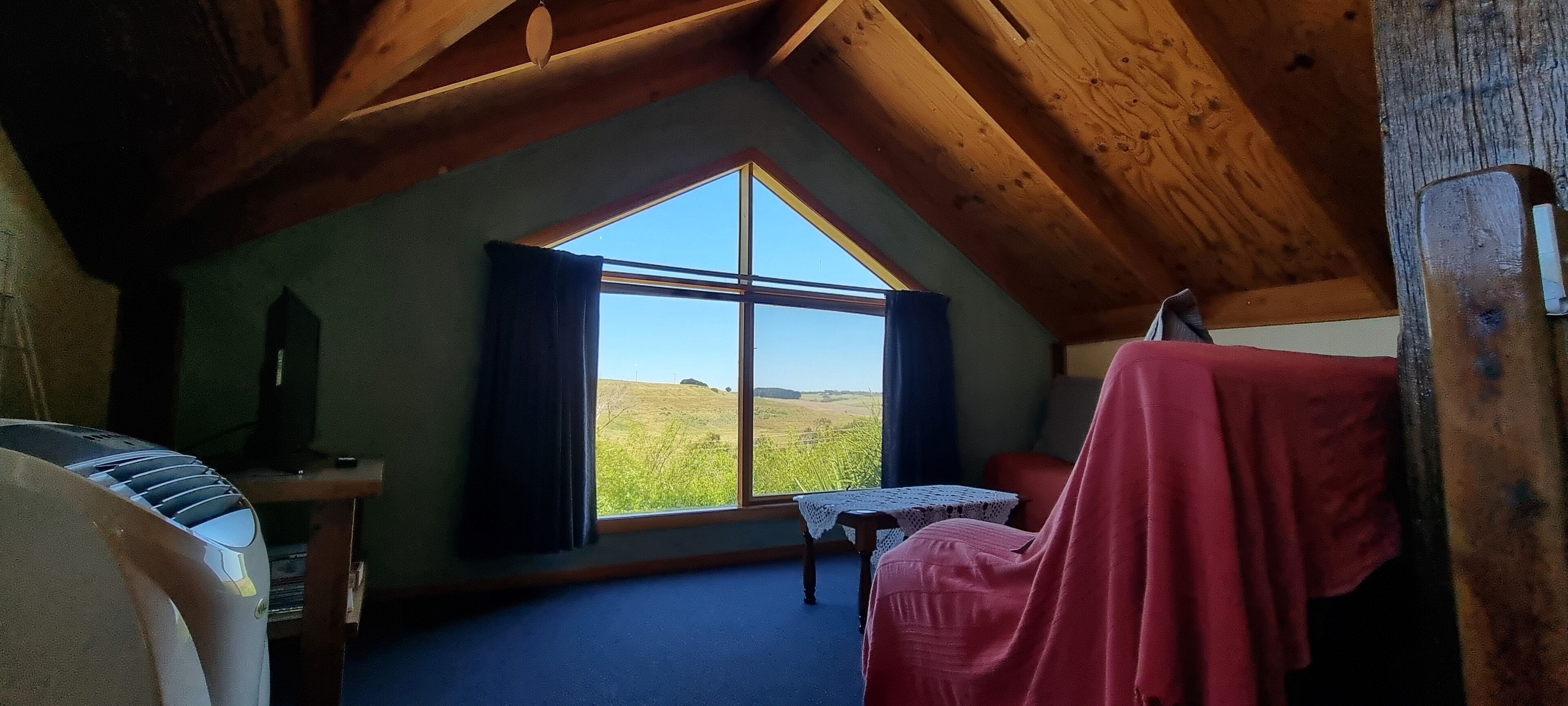 A cozy attic bedroom with a large window, wooden beams, a small table, and a chair.
