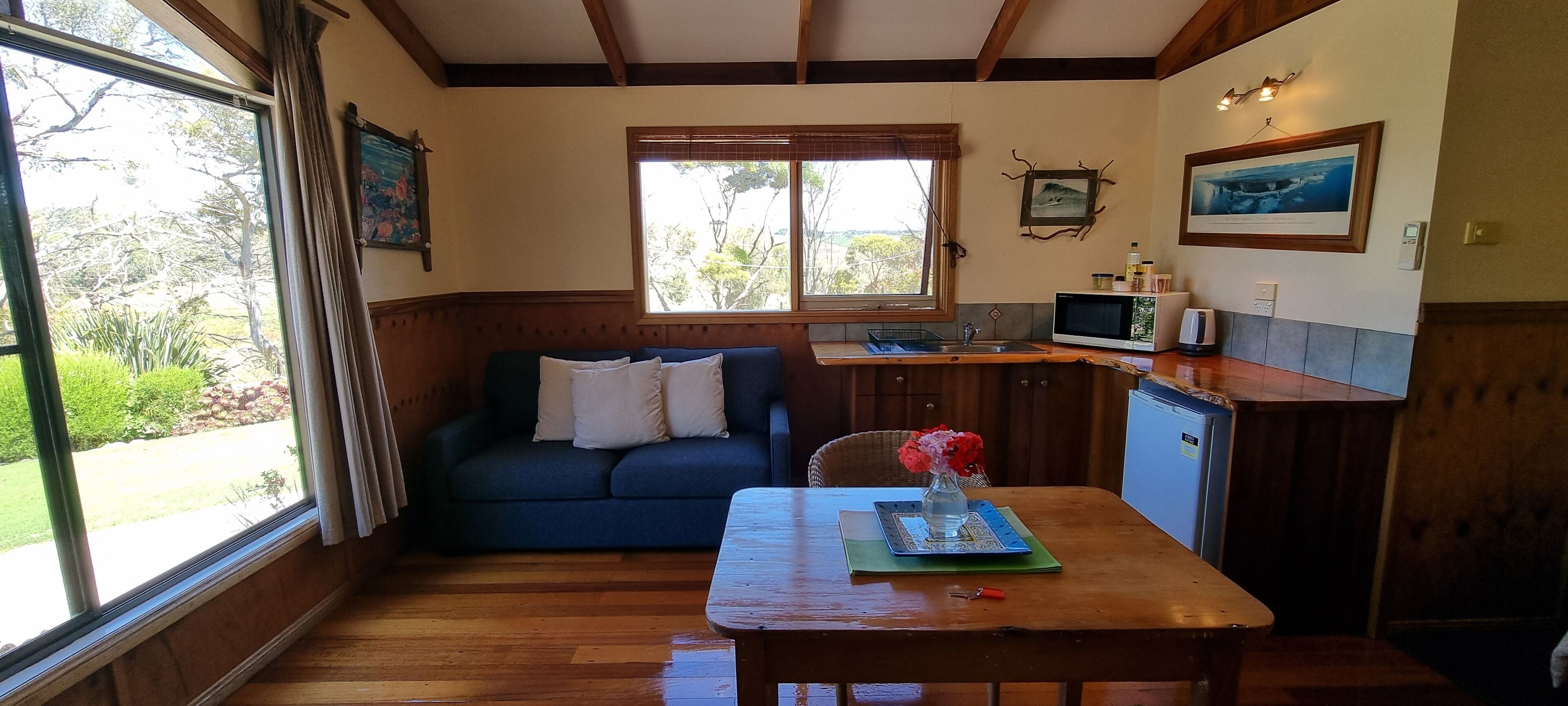 A cozy room with a wooden floor, a blue sofa, a dining table with a vase of flowers, a kitchen area with a microwave, and a window with a view of greenery.