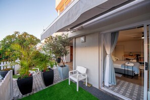 Un apartamento moderno con balcón, un pequeño jardín con macetas, una silla blanca y una vista parcial del interior con una cama y una mesita.