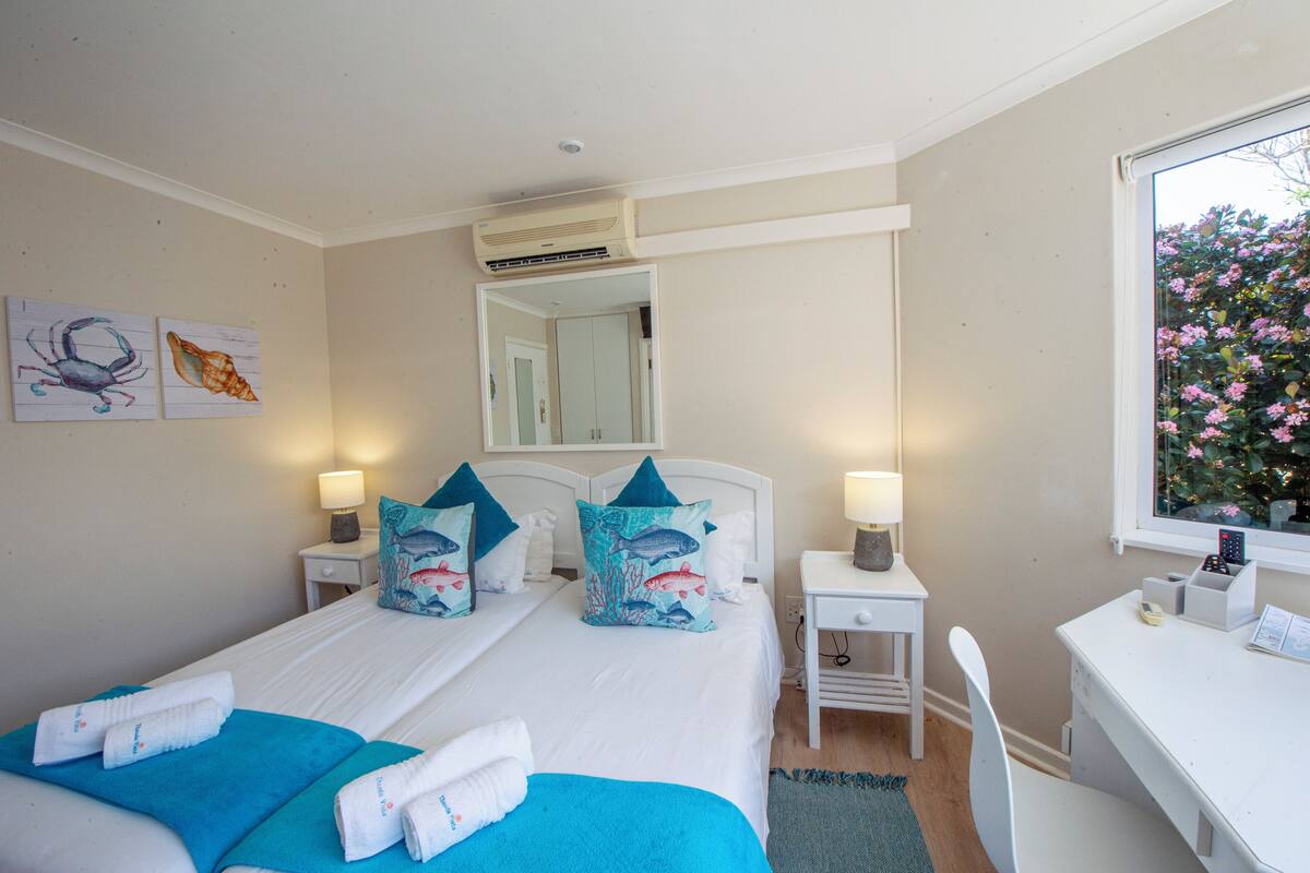 A hotel room with a bed, two bedside tables, a desk, and a window with a view of flowers.