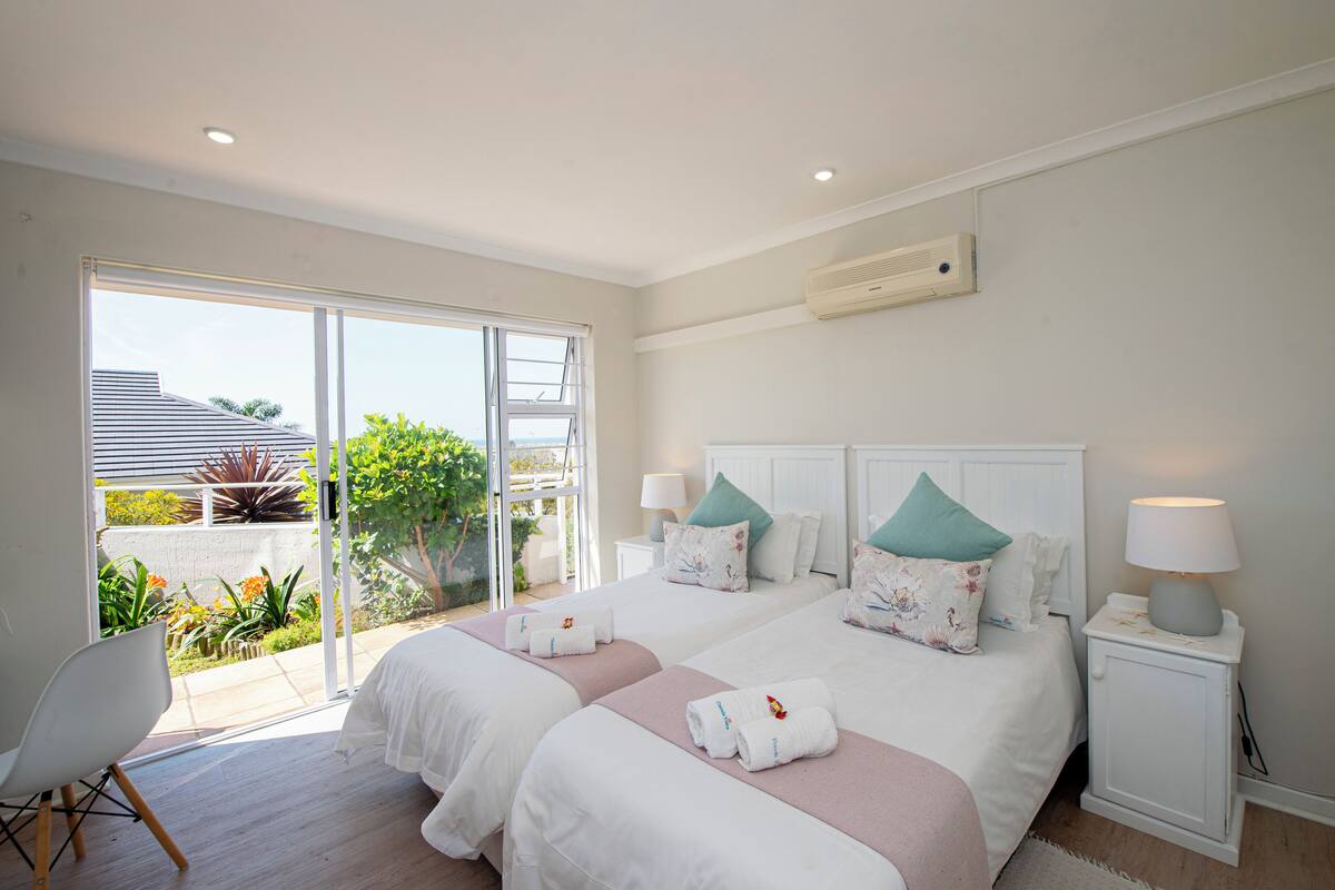 A bedroom with two beds, a sliding glass door to a balcony, a white chair, and a bedside table with a lamp.