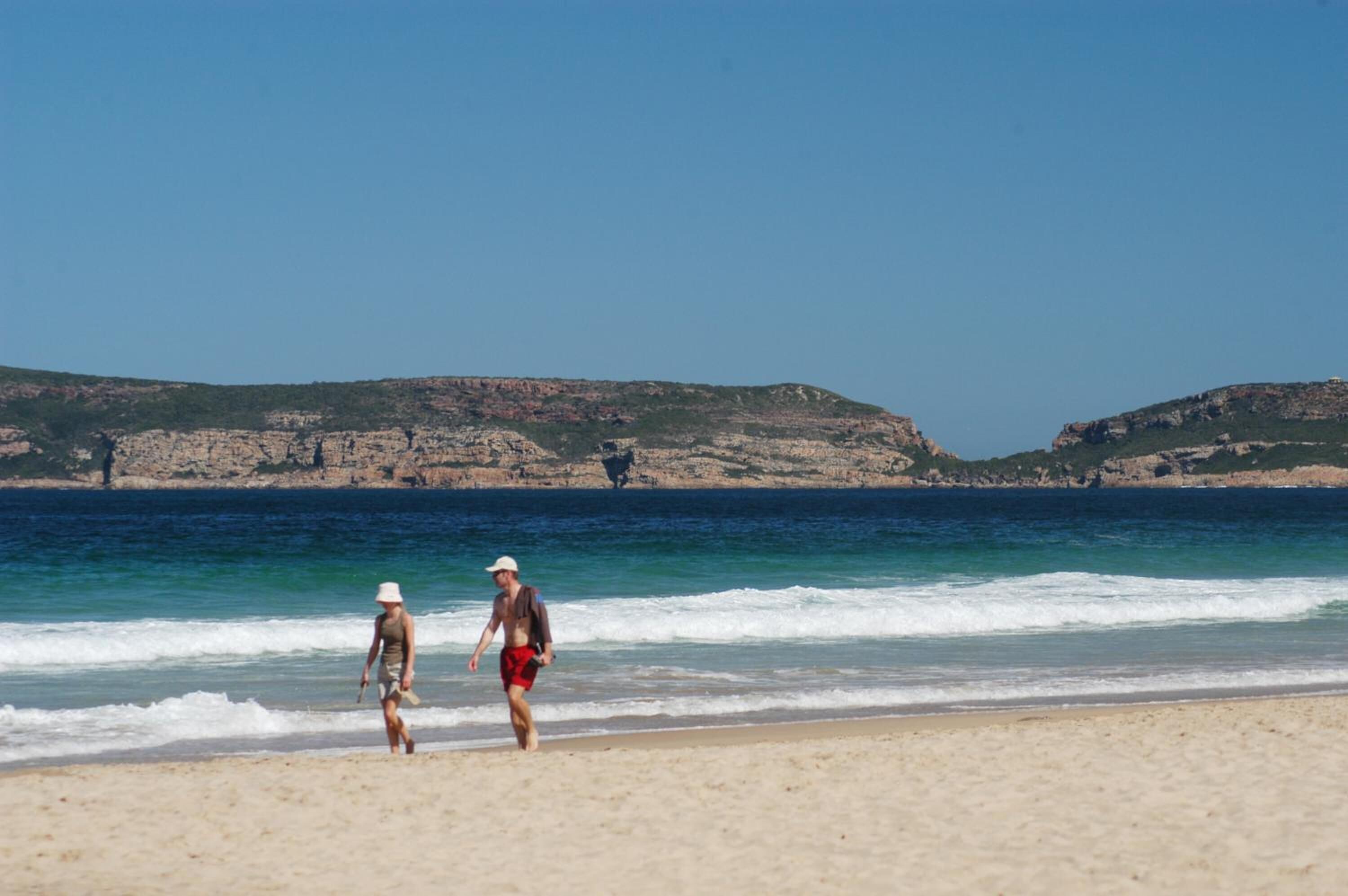 Two people walking on a sandy beach with a rocky coastline in the background.