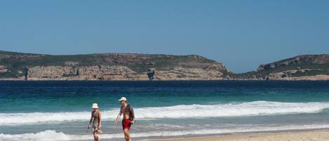 Two people walking on a sandy beach with a rocky coastline in the background.
