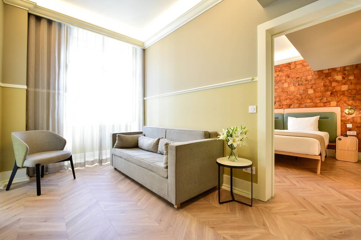 A hotel room with a sofa, a chair, a small table with a vase, a bed, and a wooden wall.